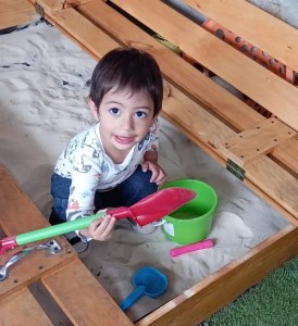 un niño juega en una caja de arena con un cubo verde y una pala roja. Está feliz
